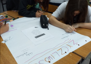 Uczennice pracują w grupie. Na stoliku jest duży arkusz papieru z napisem „Życzliwość”. Uczennice rysują plakat promujący życzliwe zachowania.